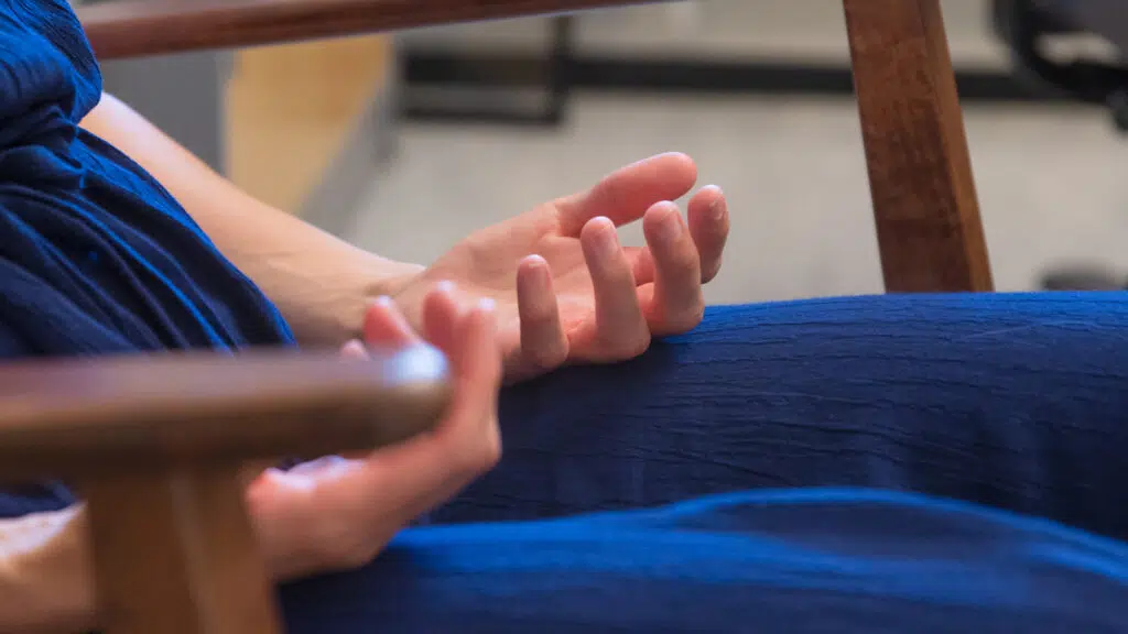 En person i blå kjole sitter med åpne hender og håndflatene vendt opp, hvilende på et armlen av tre i et innendørs miljø.