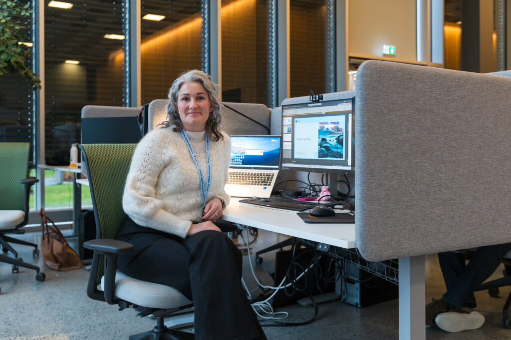 En kvinne sitter ved et skrivebord med to skjermer og en bærbar datamaskin, iført hvit genser og nøkkelbånd, i et moderne kontorlandskap med store vinduer.