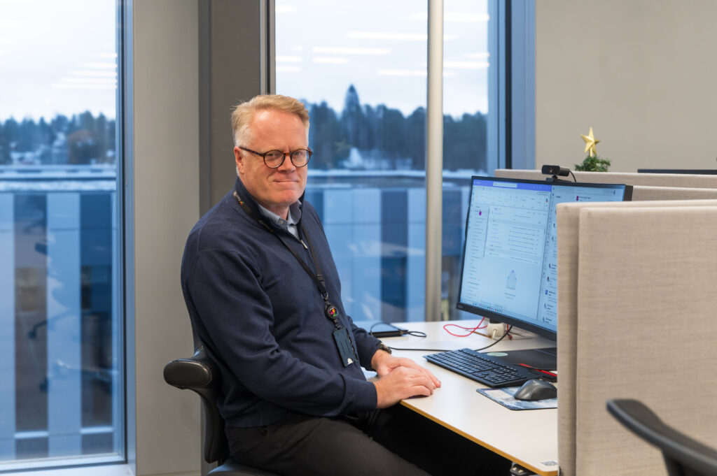 En mann med briller sitter ved et kontorpult, vendt mot to skjermer. På skilleveggen står et lite pyntet juletre, og store vinduer viser et utendørs, snødekt landskap.