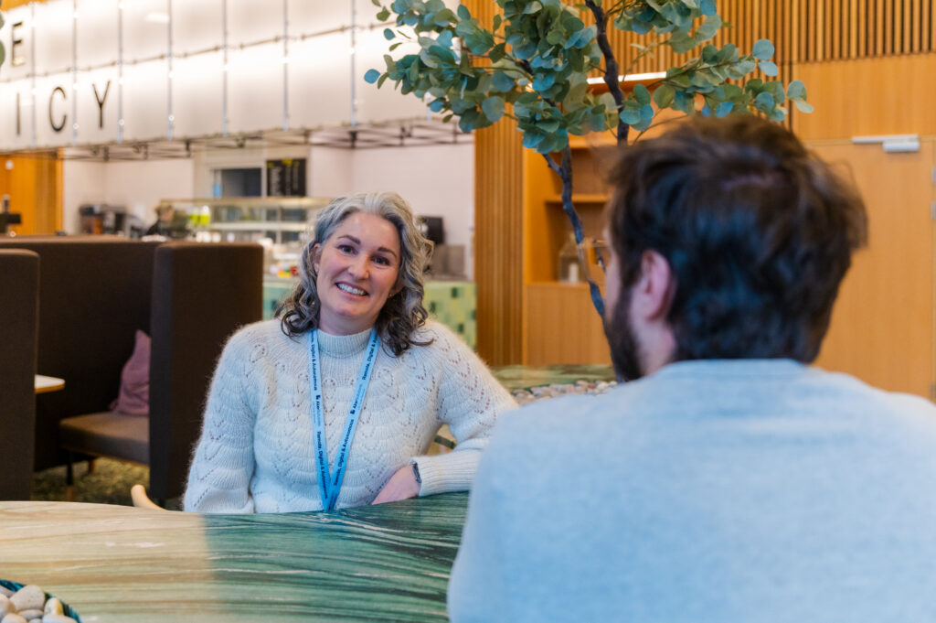 En kvinne i hvit genser og nøkkelbånd sitter ved et bord, smiler og snakker med en mann i lys genser i et moderne, innendørs miljø.