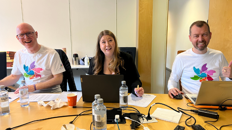 Tre personer sitter ved et konferansebord med bærbare datamaskiner og vannflasker; to av dem har matchende t-skjorter med logo, og én har en notatblokk. Alle virker engasjerte og smilende.
