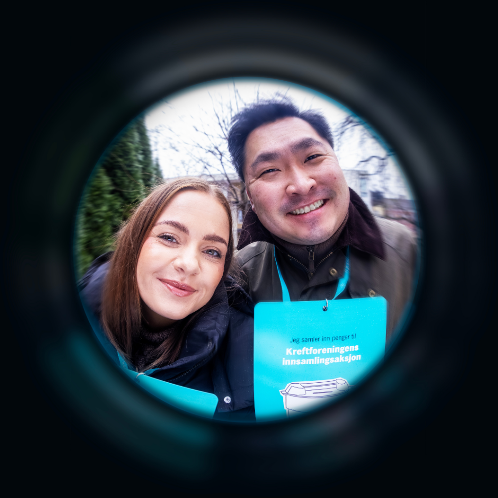 To personer med blå innsamlingsmerker smiler til kameraet utendørs, sett gjennom en sirkulær fiskeøye-effekt.