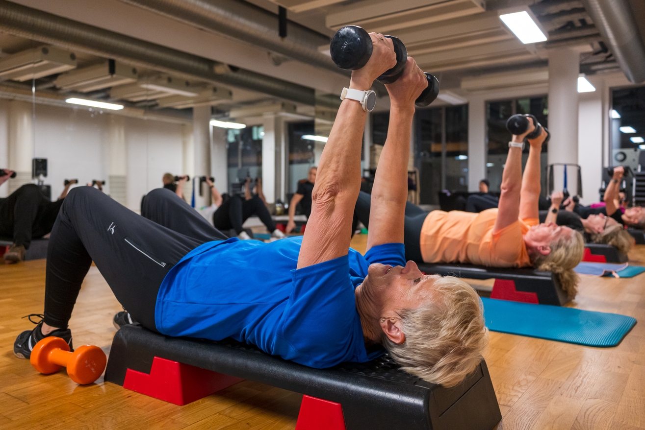 En gruppe eldre voksne ligger på benker i et treningsstudio og løfter manualer under en treningstime.