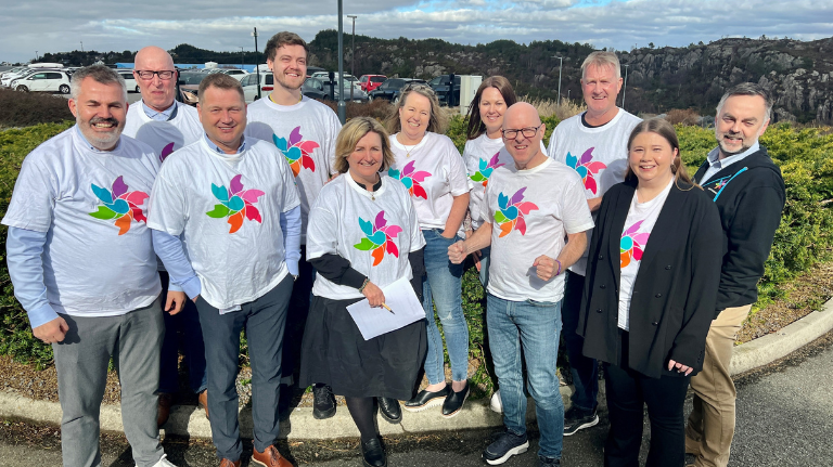 En gruppe mennesker står utendørs og smiler til kameraet. De fleste har på seg hvite t-skjorter med fargerike pinwheel-motiver. Busker og biler er synlige i bakgrunnen.