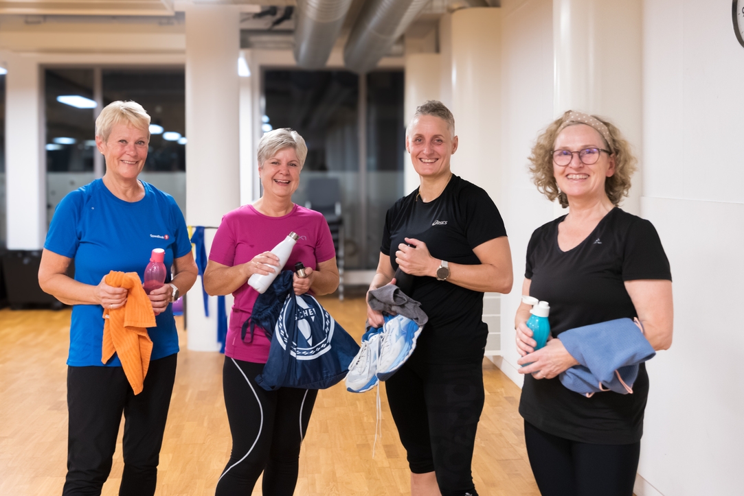 Fire voksne i treningsklær står i et treningsstudio og smiler og holder vannflasker, håndklær og sportsbager etter å ha trent.