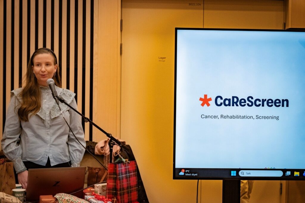 En kvinne står ved en mikrofon ved siden av en stor skjerm med CaReScreen-logoen og ordene "Cancer, Rehabilitation, Screening" i et konferanserom.