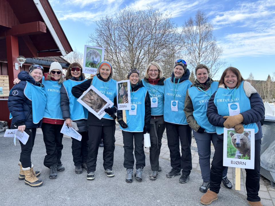 En gruppe på ni personer iført blå frivilligvester står utendørs med dyreplakater i hendene og smiler til kameraet på en solrik dag.