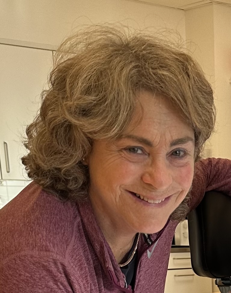 En kvinne med kort, krøllete, grått hår og en rødbrun topp smiler mens hun lener seg på en svart stol i et kjøkkenmiljø.