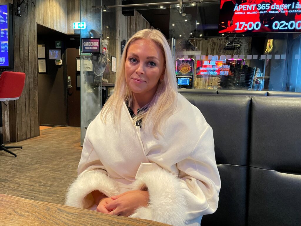 En kvinne med langt, blondt hår sitter på en svart benk i en moderne, dunkelt opplyst restaurant, iført en hvit, pelskledd frakk.