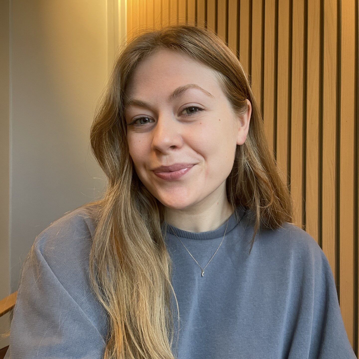 En kvinne med langt, blondt hår, iført grå genser og halskjede, sitter innendørs foran en spilevegg av tre og smiler til kameraet.