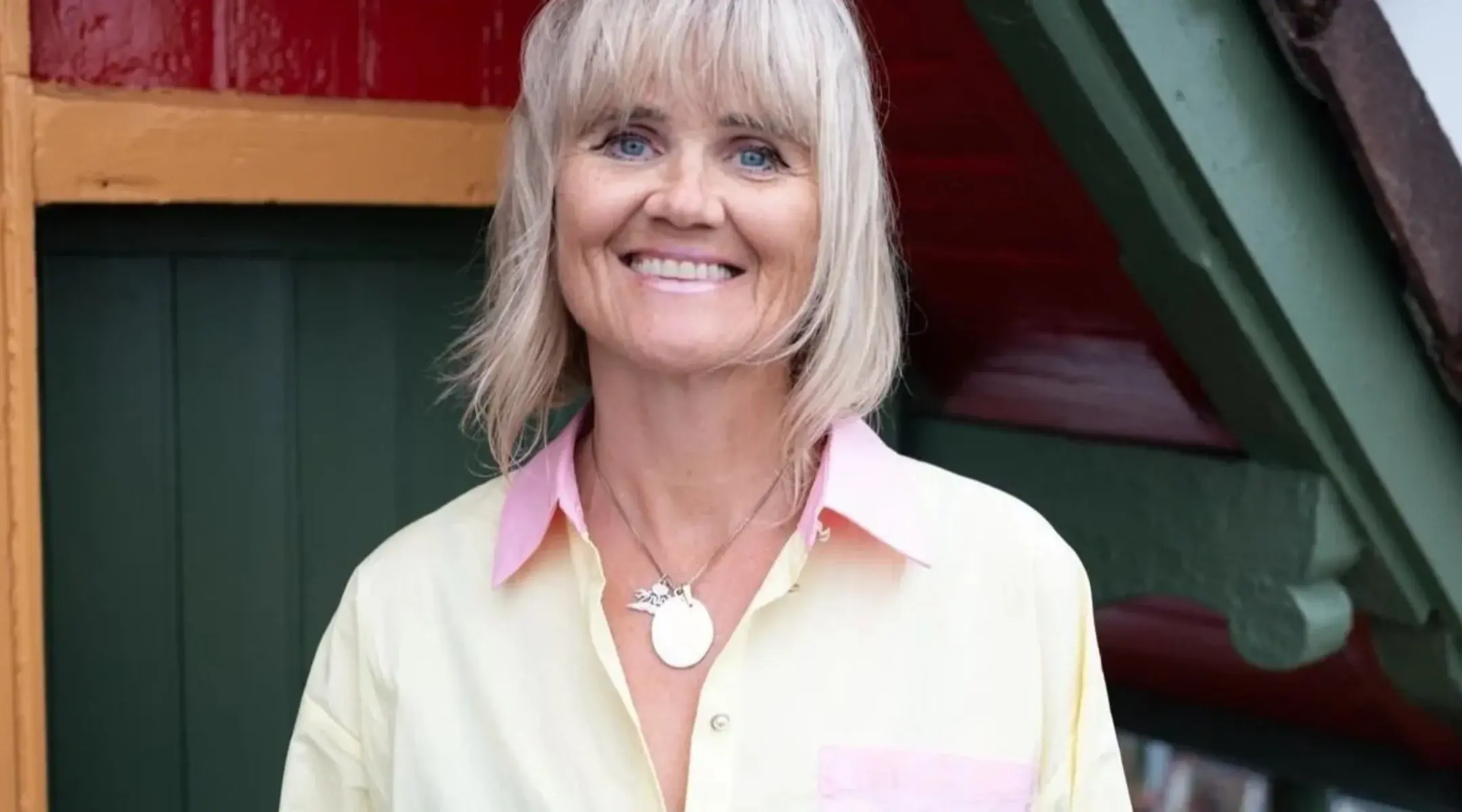 En kvinne med kort, blondt hår, iført en lysegul skjorte med rosa krage og et halskjede med anheng, smiler mens hun står foran en flerfarget bakgrunn.
