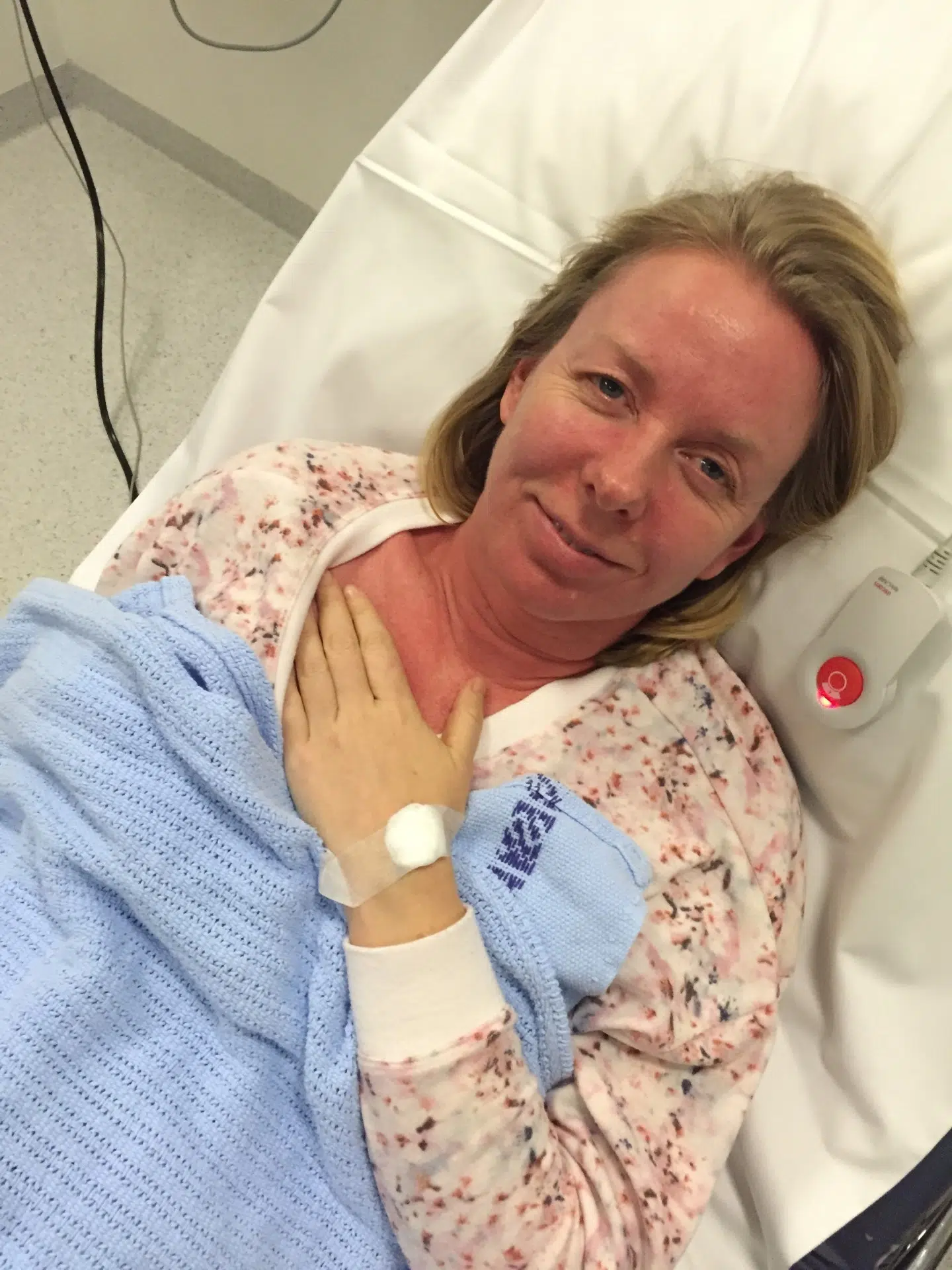 En kvinne ligger i en sykehusseng med et blått teppe, en IV-bandasje på hånden og en sykepleierknapp ved siden av seg.