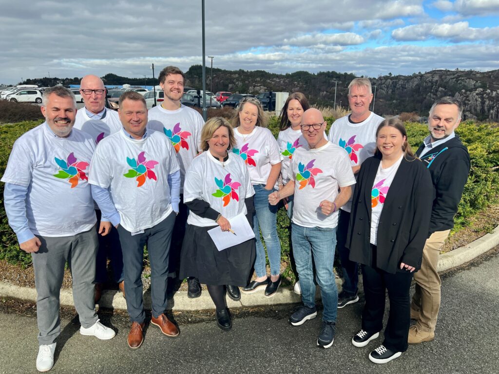 Ti voksne mennesker står utendørs, de fleste iført hvite t-skjorter med en fargerik, abstrakt logo, og poserer for et gruppebilde en delvis overskyet dag.