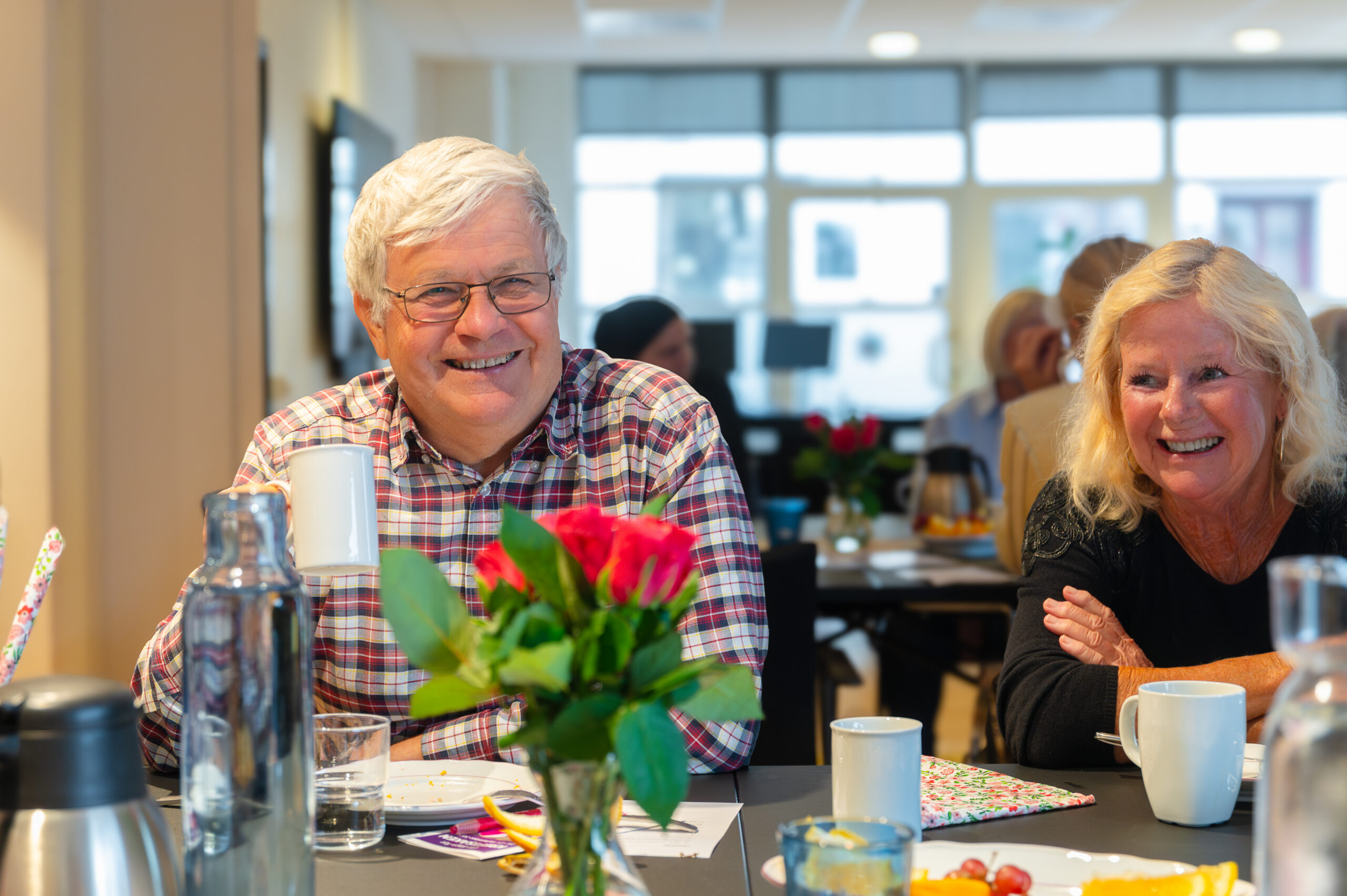 To eldre voksne sitter ved et bord med drinker og blomster, smiler og snakker sammen i et lyst rom med andre mennesker i bakgrunnen.