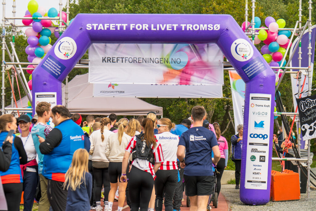 Deltakerne samles under en lilla buegang med påskriften "Stafett for Livet Tromsø" på et utendørsarrangement med ballonger, bannere og sponsorlogoer.