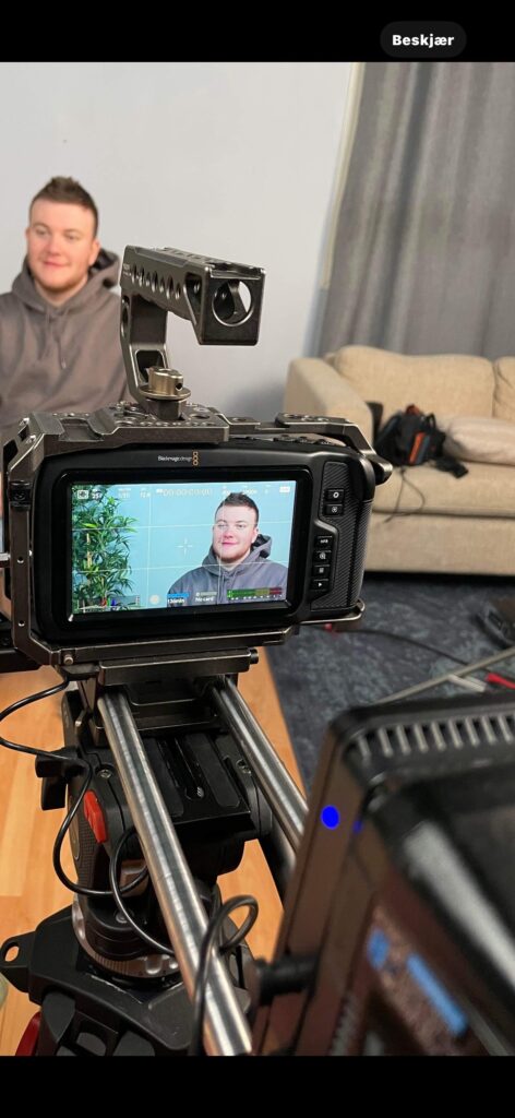 En mann sitter i et intervju med et kamera som filmer ham; bildet av ham vises på kameraets skjerm. En sofa og kamerautstyr er synlig i bakgrunnen.