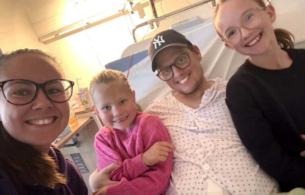 Fire personer, deriblant en sykehuspasient i kittel og lue, smiler sammen på et sykehusrom og deler håp og støtte inspirert av Golf for Livets kamp mot tarmkreft.