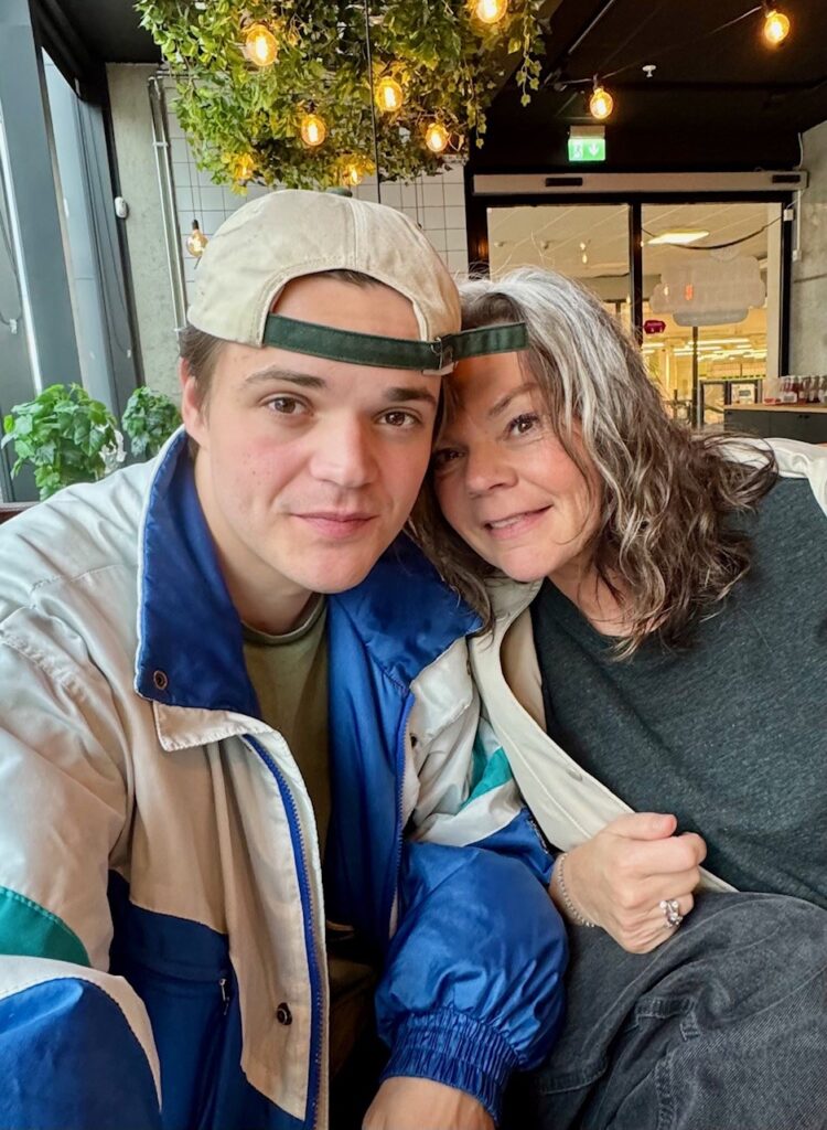 To personer sitter tett sammen innendørs. Den yngre personen har på seg caps og blå jakke, mens den eldre har langt grått hår og mørk skjorte. Begge smiler mot kameraet.