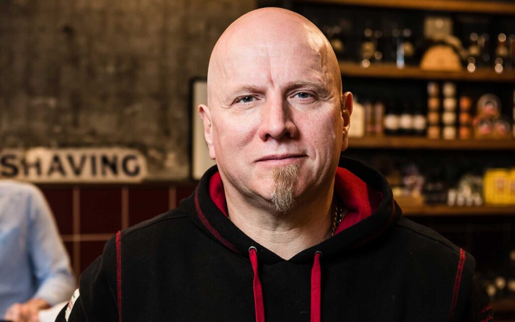 En skallet mann med fippskjegg og svart hettegenser står innendørs med hyller med pleieprodukter i bakgrunnen.