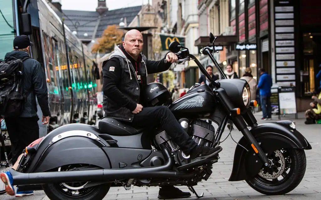 En mann sitter på en matt svart Indian-motorsykkel parkert i en bygate, med mennesker og bygninger synlige i bakgrunnen.
