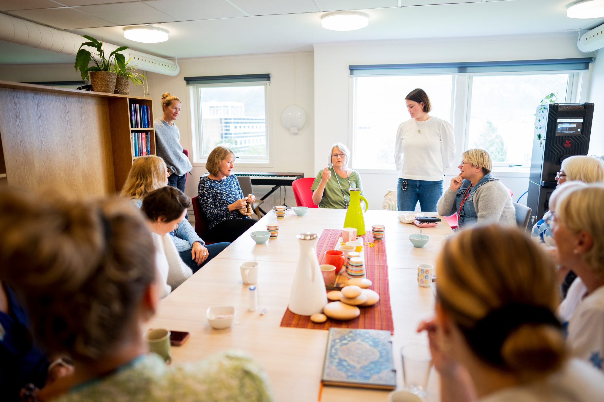 En gruppe mennesker sitter rundt et bord med drinker og snacks og snakker sammen i et lyst rom.