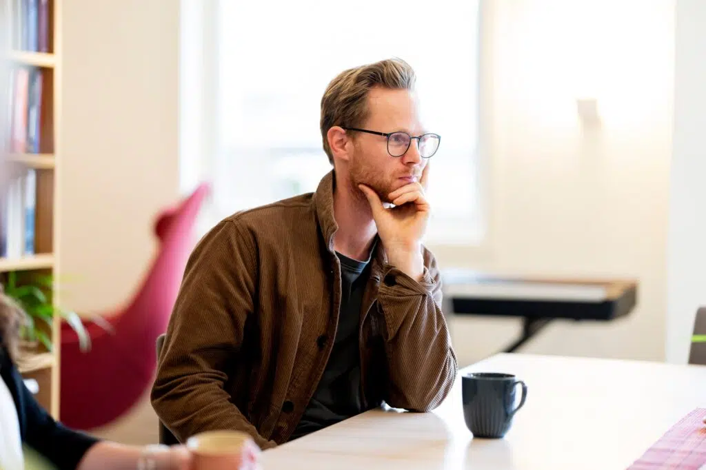 En mann med briller og brun jakke sitter ved et bord med hånden på haken og ser ettertenksom ut. Foran ham står et svart krus, og bakgrunnen er uskarp.