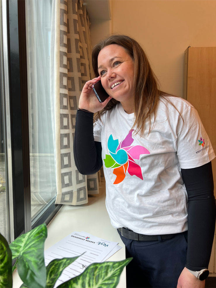 En kvinne i hvit t-skjorte med en fargerik logo smiler og snakker i telefon ved et vindu, med et dokument og en plante på bordet foran seg.