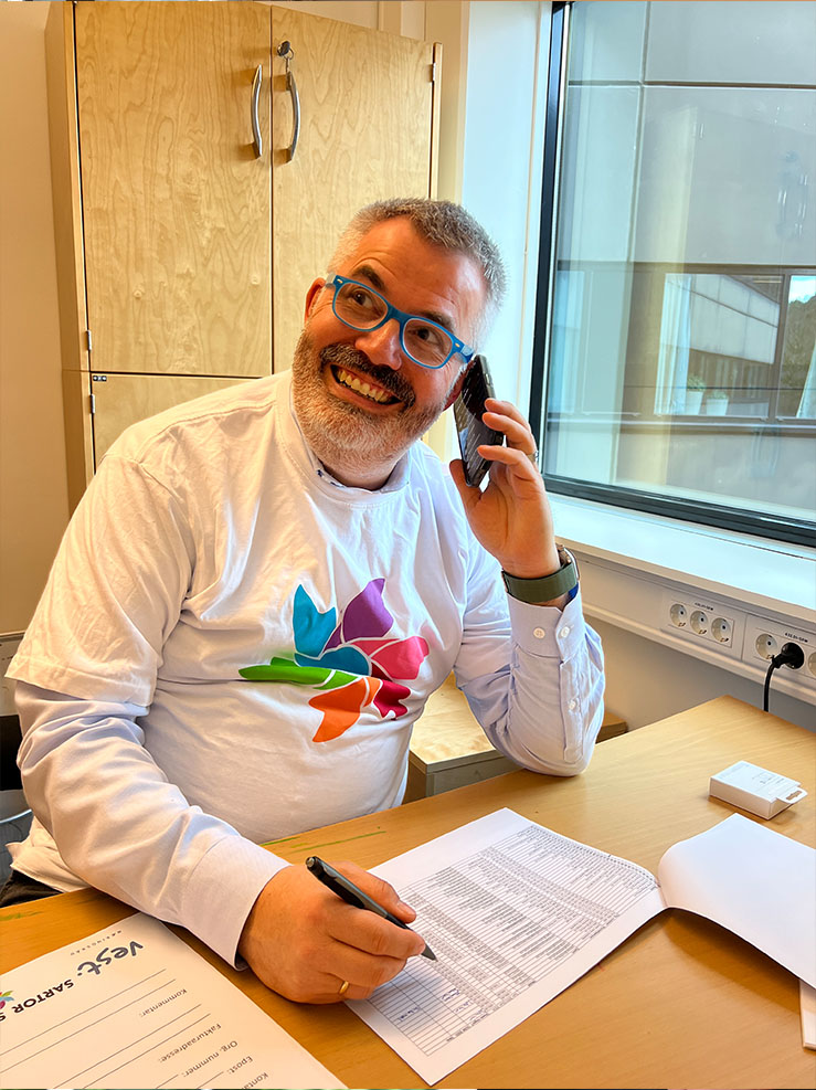 En smilende mann med briller snakker i telefon mens han sitter ved et skrivebord med en penn i hånden og går gjennom dokumenter. Han har på seg en hvit skjorte med et fargerikt, abstrakt motiv.