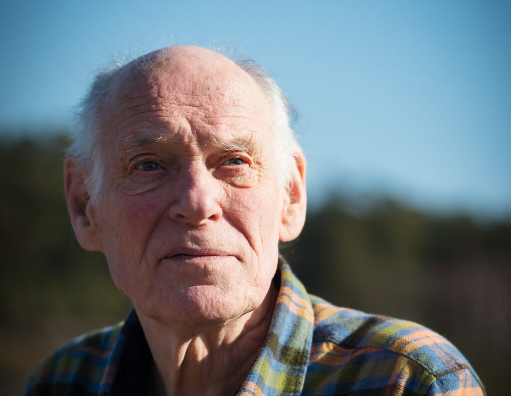 En eldre mann med tynt, hvitt hår og rutete skjorte ser ettertenksomt ut i det fjerne en solfylt dag utendørs.