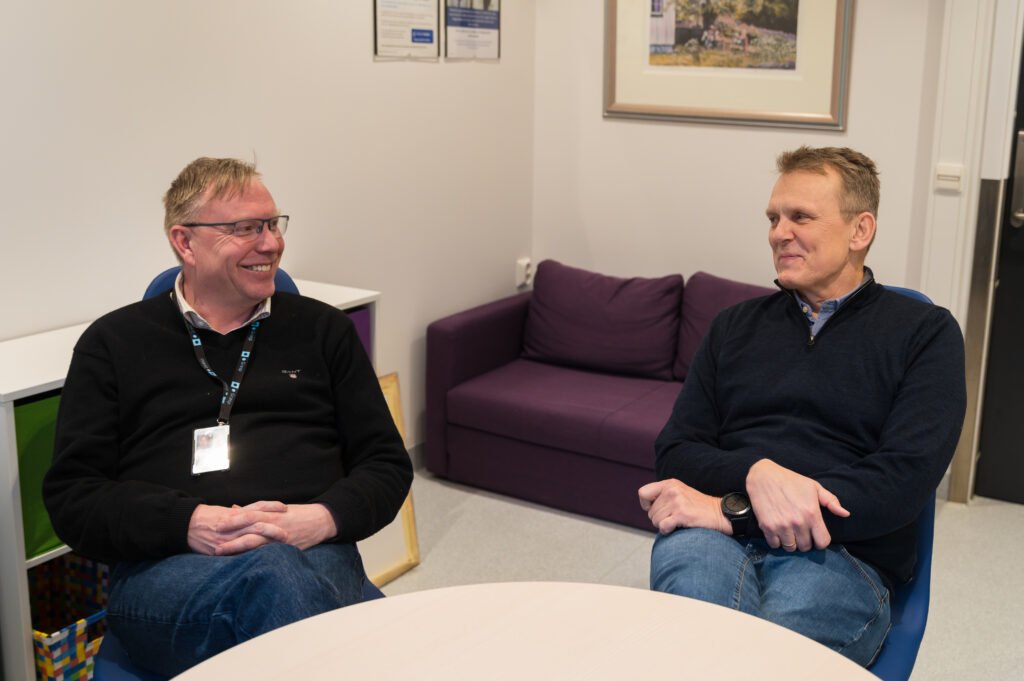 To menn sitter og snakker ved et rundt bord i et kontormiljø, begge iført briller og mørke gensere, med en lilla sofa og et innrammet bilde i bakgrunnen.