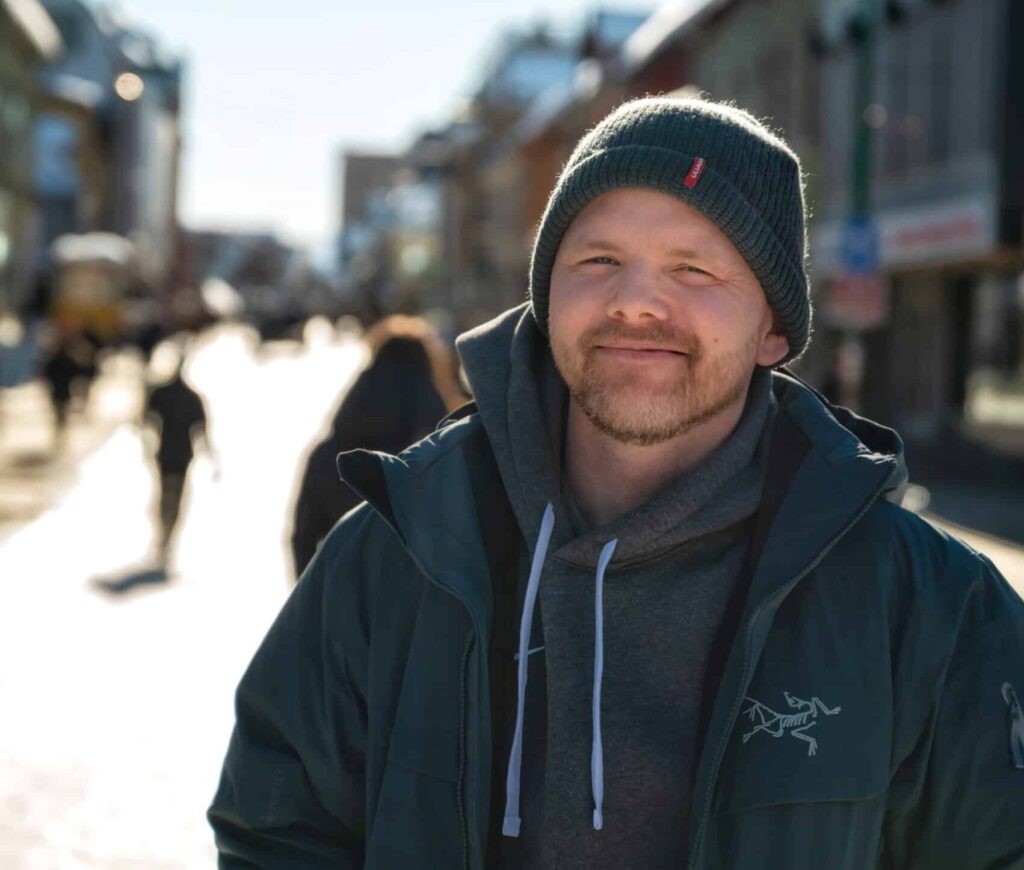 En mann i lue og jakke smiler utendørs på en solrik dag, stående i en bygate med uskarpe fotgjengere i bakgrunnen.