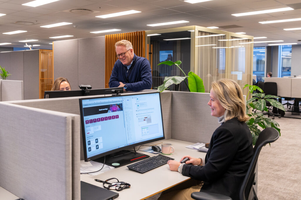 Tre personer i et moderne kontorlandskap, to sitter ved pulter med datamaskiner og én står og snakker sammen. Arbeidsområdet har avgrensede avlukker og inneplanter.