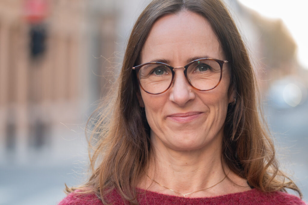 En kvinne med langt brunt hår og briller står utendørs og smiler lett, iført rød genser og et fint halskjede. Bakgrunnen er mykt uskarp.
