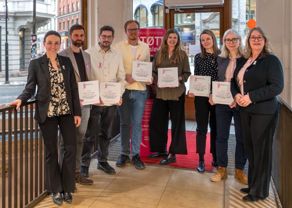 Åtte personer står innendørs med diplomer og rosa sløyfepins og poserer for et gruppebilde i nærheten av et vindu og et rosa skilt.