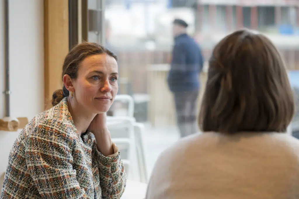 To kvinner sitter overfor hverandre ved et bord ved et vindu, i samtale. Den ene kvinnen er vendt mot kameraet, mens den andre har ryggen til.