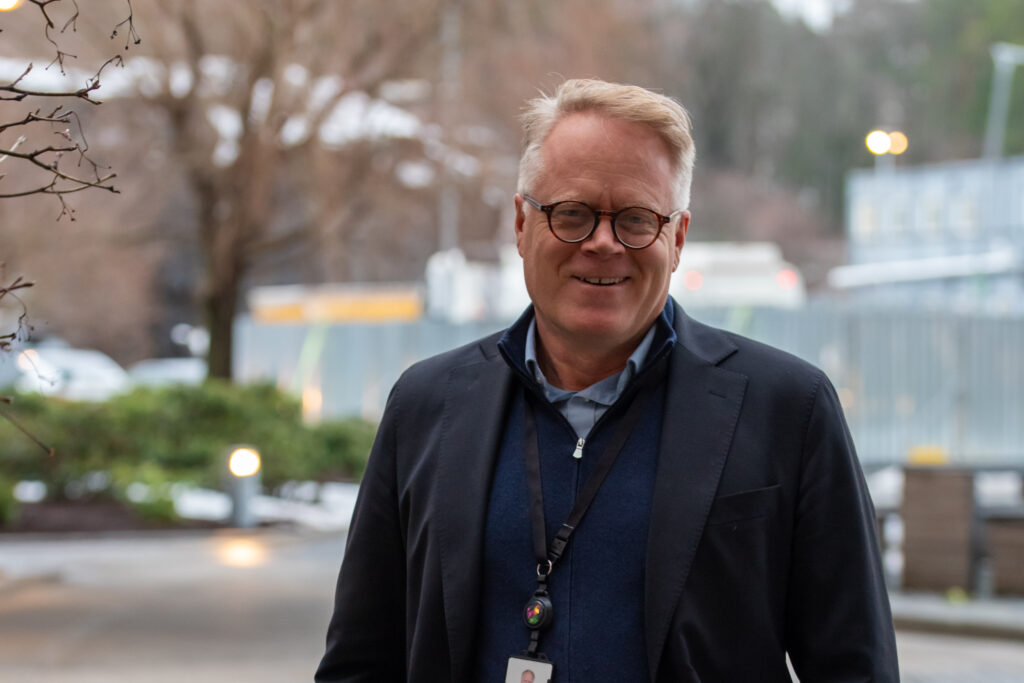 En mann med briller og mørk jakke står utendørs og smiler til kameraet, med trær og uskarpe bygninger i bakgrunnen.