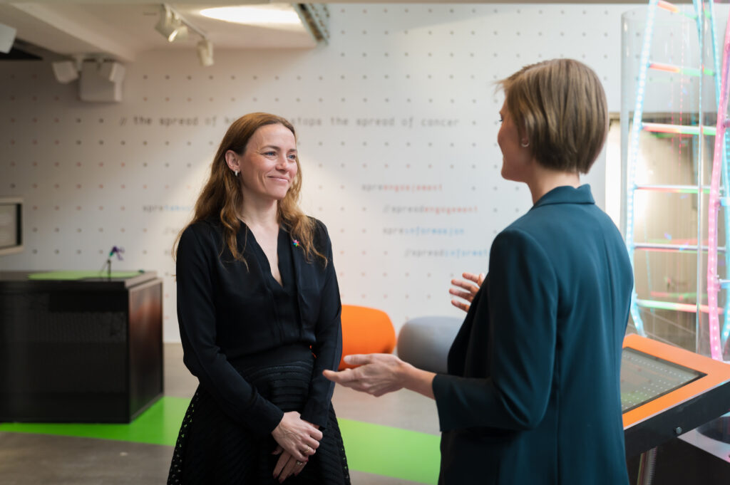 To kvinner står og snakker sammen i et moderne, sterkt opplyst kontor- eller utstillingslokale.