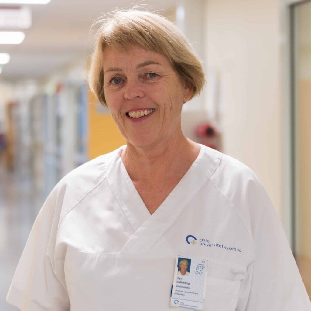 En kvinne i hvit legeuniform med logo fra Oslo universitetssykehus står i en gang og smiler til kameraet. Hun har et navneskilt festet til skjorten.