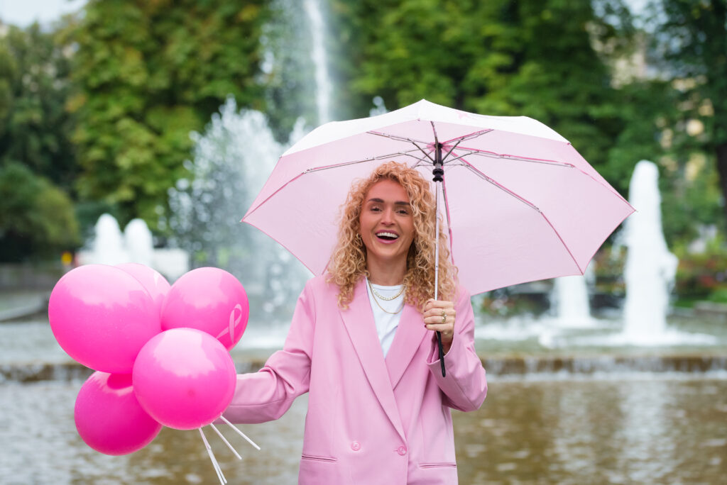 En person i rosa blazer holder rosa ballonger og en rosa paraply foran en fontene i en park.