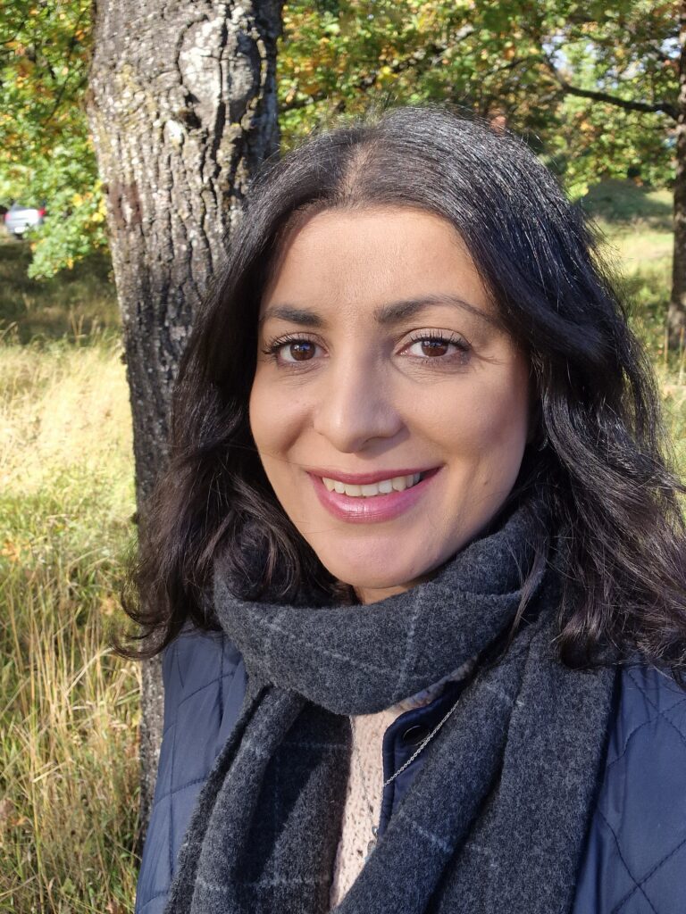 En kvinne med mørkt hår og grått skjerf smiler utendørs på en gresslette med trær og sollys i bakgrunnen.