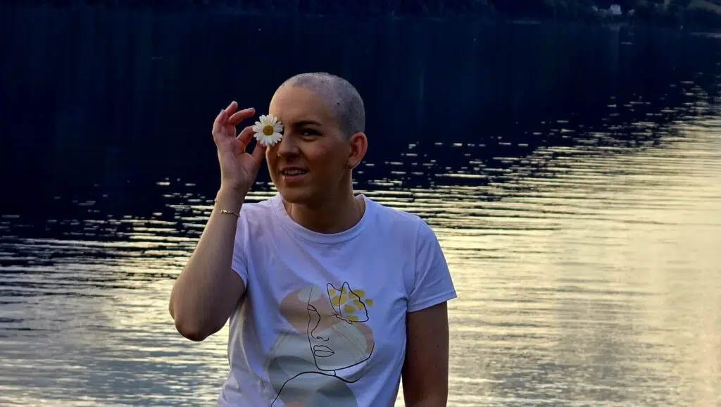 En person med barbert hode holder en tusenfryd over det ene øyet, smiler og står foran en rolig innsjø i solnedgang iført en hvit, grafisk t-skjorte.