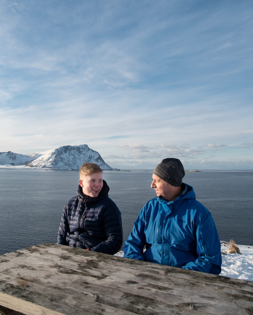To personer i vinterjakker sitter ved et trebord utendørs med snødekte fjell og et stort vann i bakgrunnen.