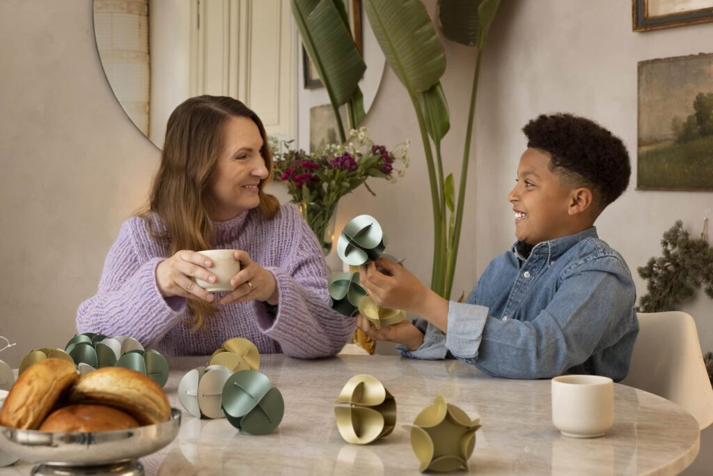 En kvinne og et barn sitter ved et bord med bakverk, drikke og pyntegjenstander, og de smiler og snakker sammen.