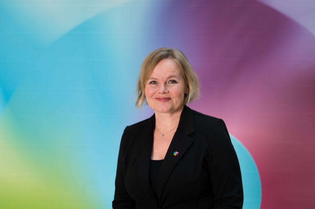 En kvinne i svart blazer står foran en fargerik, tonet bakgrunn og smiler til kameraet.