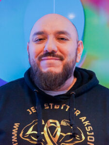 En skjeggete mann i svart hettegenser står foran en fargerik, uskarp bakgrunn, ser inn i kameraet og smiler mildt.