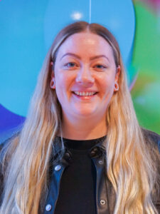 En kvinne med langt, blondt hår og utstrakte øreflipper smiler til kameraet, iført svart skjorte og svart skinnjakke, med fargerike ballonger i bakgrunnen.