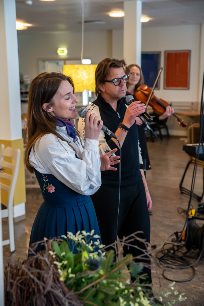 En kvinne synger i en mikrofon mens en mann spiller fiolin i bakgrunnen på et innendørsarrangement.