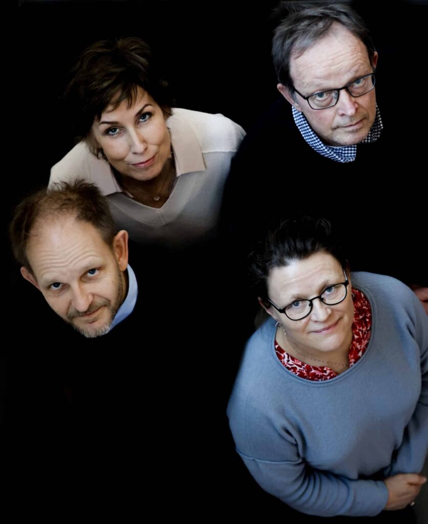 Fire voksne, to menn og to kvinner, ser opp mot kameraet og står tett sammen mot en mørk bakgrunn.