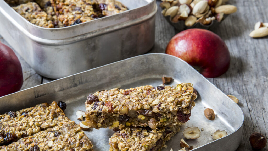 Hjemmelagde granolabarer med nøtter og tørket frukt i et metallbrett, med hele nøtter og røde epler på et underlag av tre.