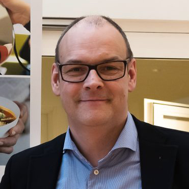 En middelaldrende mann med briller, kort hår, blå skjorte med krage og mørk blazer står innendørs og smiler til kameraet.